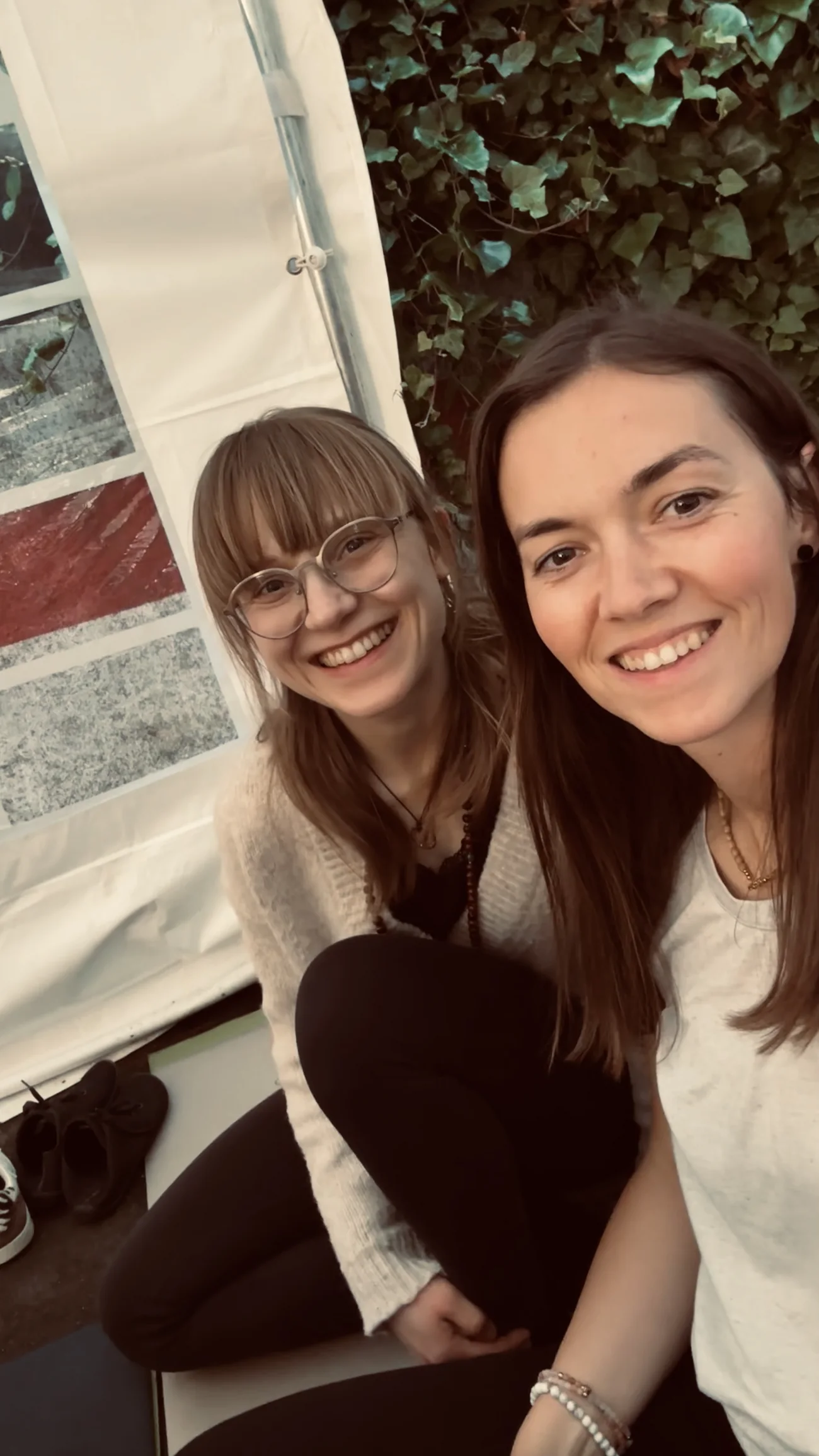 Alicia und Laura Schadt sitzen nebeneinander und lächeln in die Kamera, im Hintergrund eine Efeuwand.