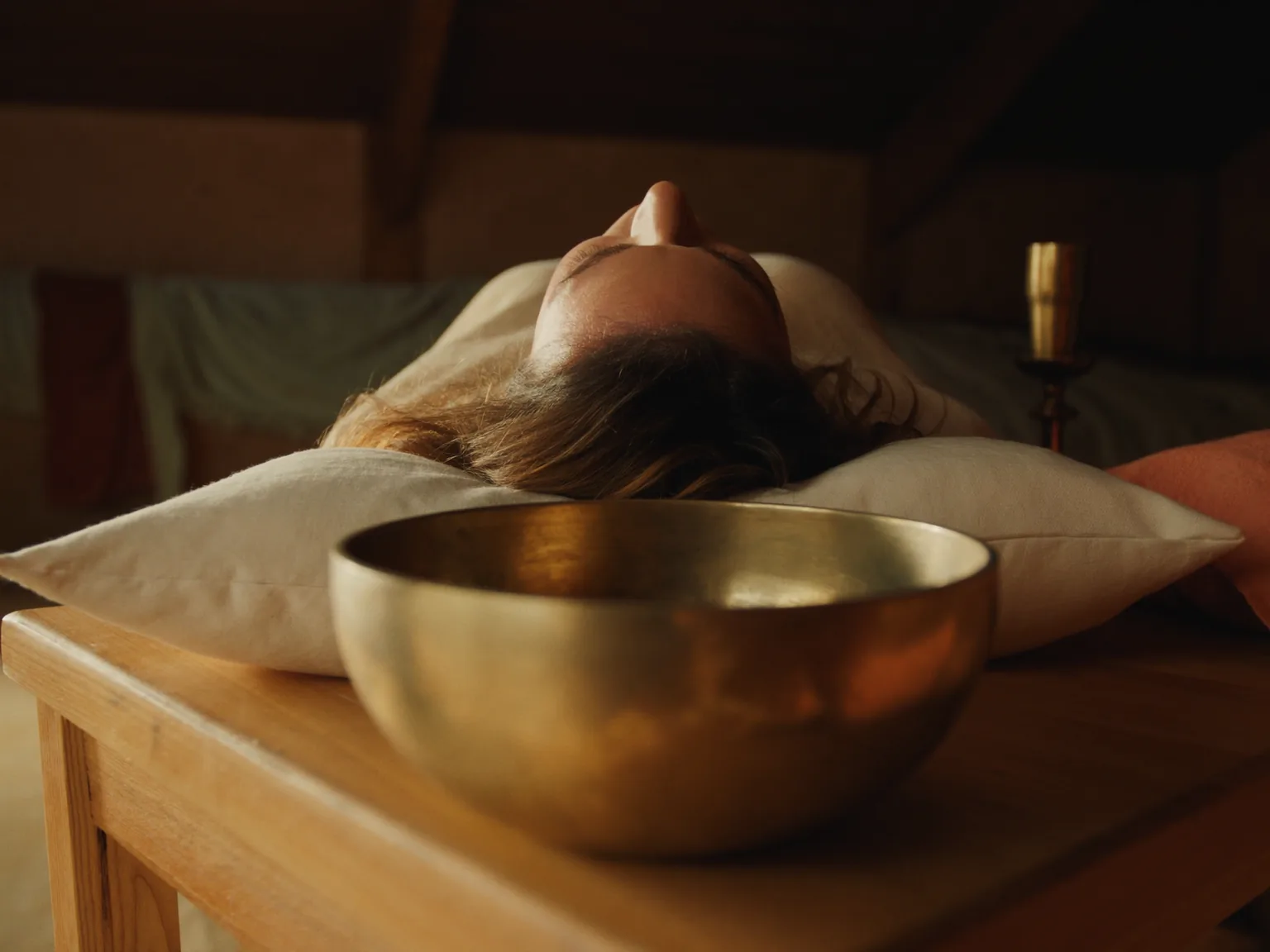 Klangmassage-Sitzung: Blick von hinter einer goldenen Messing-Klangschale, die am Kopfende einer entspannt liegenden Frau steht. Die Frau hat geschlossene Augen, ruhiger Gesichtsausdruck, liegt auf einer hölzernen Massageliege im warmen, in Holztönen gehaltenen Raum.